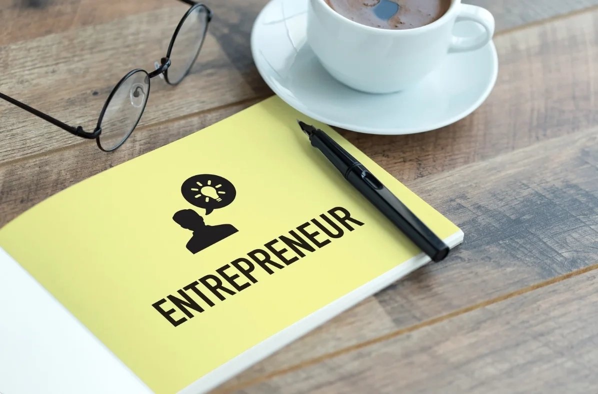 Esprit entrepreneurial – Accompagnement BY « Carnet jaune marqué entrepreneur posé sur un bureau avec lunettes, stylo et tasse de café, symbolisant la créativité et la stratégie entrepreneuriale. »